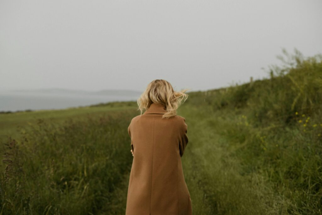 A woman in a coat walking alone on a grassy pathway by the sea under a gloomy sky.