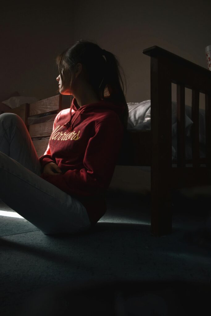 Contemplative young woman in hoodie sitting on the floor in dimly lit room, symbolizing solitude and introspection.